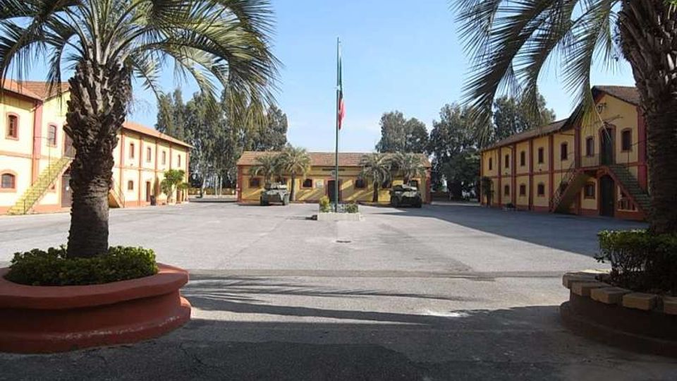 Ampio cortile pavimentato circondato da edifici bassi color ocra con finestre regolari e scale esterne, con palme in grandi fioriere ai lati. Al centro si trova un pennone con bandiera e sullo sfondo si vedono alberi oltre gli edifici.