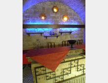Interno in pietra con volta a botte e parete di blocchi illuminata da luci blu e tre applique circolari. In primo piano un bancone in muratura con inserti in ferro battuto e tovaglia rossa, con sedie e mensole in legno sullo sfondo.