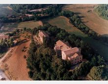 Vista aerea di una tenuta rurale con edifici in muratura dai tetti in coppi, circondati da alberi e campi coltivati. Intorno si estendono appezzamenti agricoli e strade di campagna che attraversano la campagna ondulata.