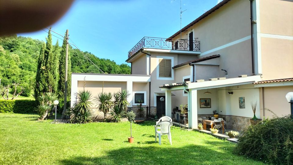 Casa di campagna a due piani con facciata chiara, balcone con ringhiera metallica e portico coperto arredato. Davanti c’è un ampio prato verde con piante ornamentali e lampioni da giardino, sullo sfondo colline boscose e cielo azzurro.