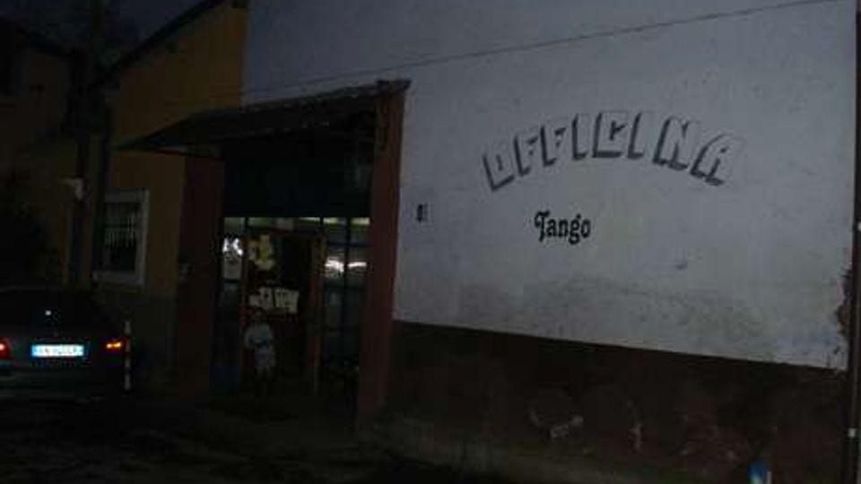 Facciata esterna di un edificio con intonaco chiaro e basamento scuro, con un grande ingresso ad arco che conduce a un’area interna illuminata. Sulla parete è visibile una scritta in rilievo e la parola “Mango”, in un contesto urbano notturno.