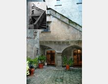 Cortile interno di un edificio storico in pietra e intonaco, con scala esterna in muratura che sale lungo la parete e un porticato ad archi con colonne. Sul pavimento in mattoni a spina di pesce ci sono grandi vasi con piante, e sullo sfondo si vedono porte con grate in ferro battuto.