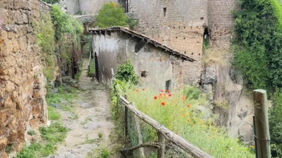 Sentiero in pietra in discesa tra alte mura di un complesso fortificato in rovina, con una torre cilindrica sullo sfondo. Ai lati si vedono una staccionata in legno e vegetazione spontanea, con un piccolo edificio addossato alle mura sotto un cielo nuvoloso.