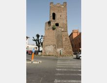 Torre medievale in mattoni con merlature e aperture ad arco, affiancata da resti di muratura e base scarpata. Si trova su un incrocio urbano con strisce pedonali, segnaletica stradale, alberi e palazzi moderni sullo sfondo.