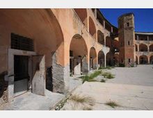 Cortile interno di un complesso storico in rovina, con porticati ad archi su più livelli e intonaco scrostato. Ampio pavimento in cemento con ciuffi d’erba, porte e aperture murate lungo le arcate e una torre in muratura sullo sfondo sotto cielo sereno.
