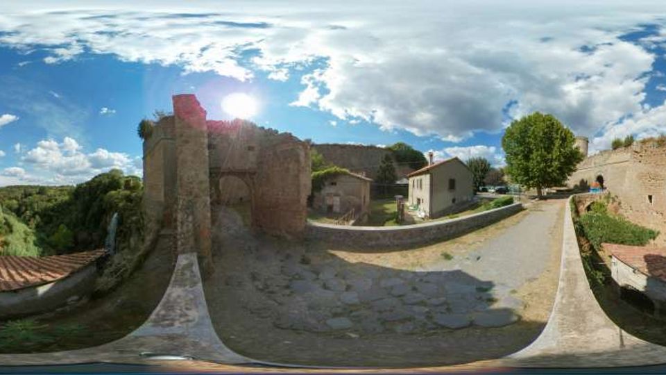 Panorama a 360 gradi di un complesso fortificato in rovina con mura e torri in pietra, affacciato su una valle alberata. Un camminamento in pietra con parapetti conduce tra edifici bassi e resti delle mura, con un grande albero e cielo parzialmente nuvoloso sullo sfondo.