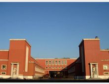 Edificio istituzionale in mattoni rossi con ali simmetriche e due torri laterali, caratterizzato da finestre rettangolari in file regolari. All’ingresso, nicchie con statue e un passaggio centrale verso il cortile interno sotto un cielo sereno.