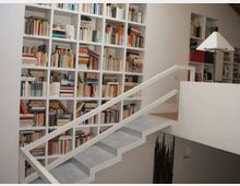 Interno di una casa con grande libreria a parete composta da scaffali bianchi a griglia pieni di libri. Una scala moderna bianca con ringhiera attraversa l’immagine e collega a un soppalco con parapetto.