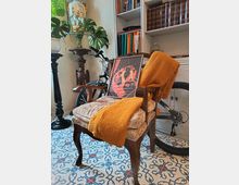 Interno domestico con poltrona in legno e seduta imbottita, coperta arancione e quadro appoggiato. Sullo sfondo scaffali bianchi con libri, una bicicletta, piante e lampade, su pavimento in piastrelle decorative blu e bianche.