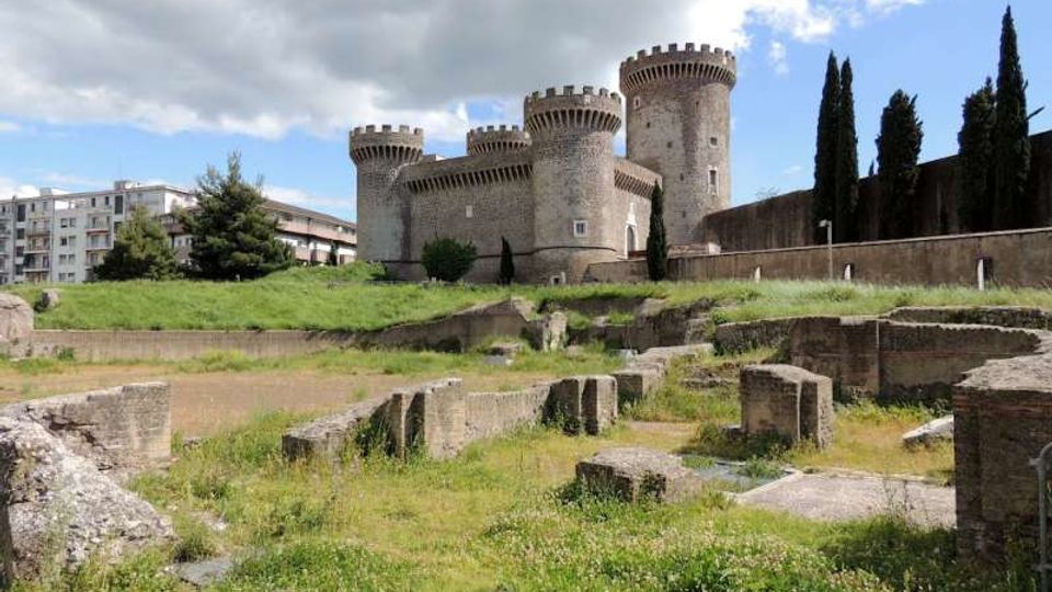 Castello medievale in pietra con quattro torri cilindriche merlate, visto da un’area archeologica con muretti e resti in muratura tra erba e sentieri. Sullo sfondo si notano cipressi e alcuni edifici moderni sotto un cielo parzialmente nuvoloso.