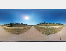 Vista panoramica di un ampio viale sterrato in un’area archeologica, con prati secchi ai lati e colline basse. Sullo sfondo si vedono rovine in muratura, filari di cipressi e un cielo azzurro con il sole alto.