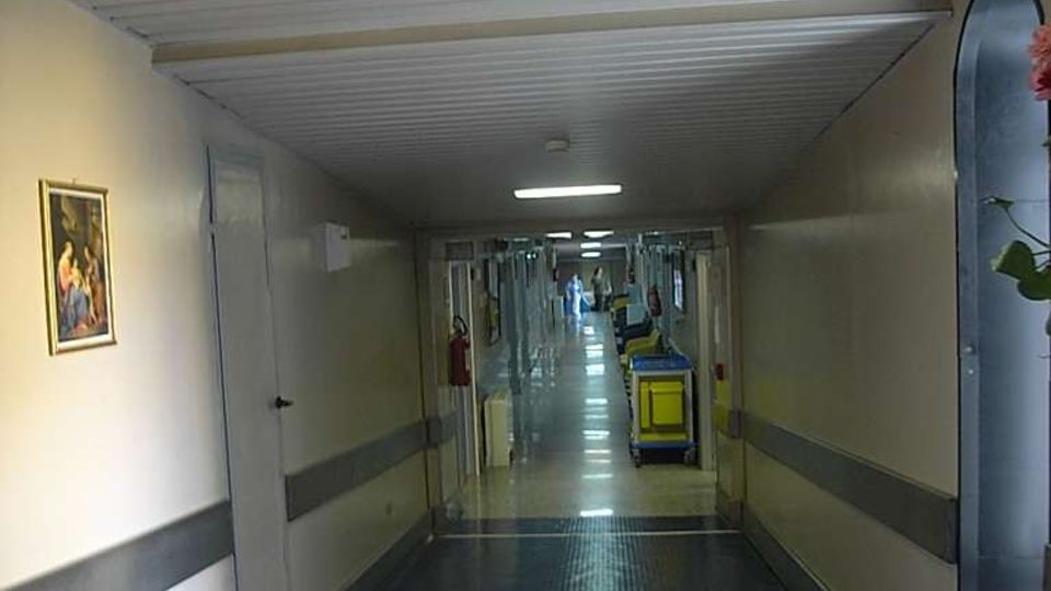 Lungo corridoio interno di un ospedale con pavimento blu lucido, pareti chiare con fascia grigia e illuminazione a plafoniere rettangolari sul soffitto. In fondo si vedono carrelli di servizio e porte lungo i lati del corridoio.