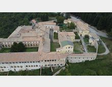 Veduta aerea di un grande complesso monastico o abbaziale con edifici in pietra e intonaco, tetti in coppi e cortili interni, con una chiesa e un campanile al centro. Il complesso è circondato da boschi e prati, con una strada di accesso sul lato destro.