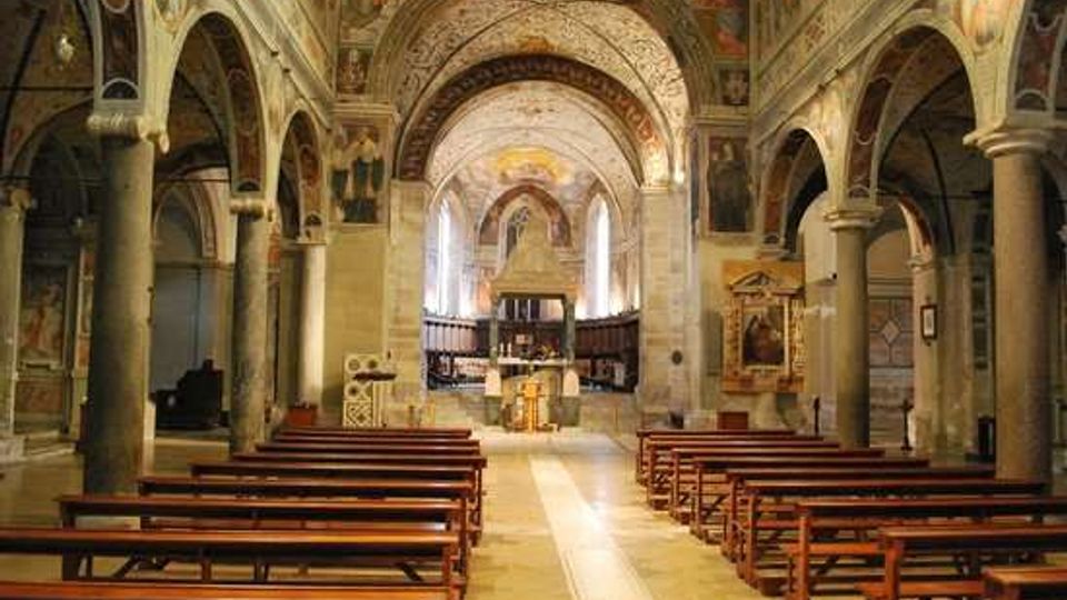 Interno di una chiesa in stile romanico con navata centrale e arcate su colonne in pietra, soffitti e pareti decorati con affreschi. Panche di legno disposte su entrambi i lati conducono verso l’altare in fondo, illuminato da una luce proveniente dall’abside.