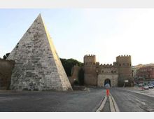 Piazza con ampia piramide rivestita in pietra in primo piano e, sullo sfondo, una fortificazione in mattoni con mura merlate, due torri e un grande arco d’ingresso. Strada asfaltata e area urbana circostante visibili ai lati.