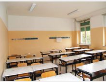 Aula scolastica con banchi e sedie disposti in file, pareti bicolori (bianco e beige) e lampade fluorescenti a soffitto. Sul lato destro ci sono grandi finestre con tapparelle verdi che illuminano la stanza.
