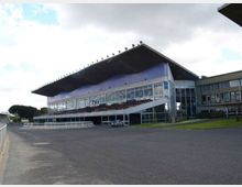 Vista esterna di una tribuna di ippodromo con grande tetto sporgente e facciata vetrata su più livelli, con rampa laterale e ingresso al piano terra. Davanti si estende una vasta area asfaltata, con prato e alcuni edifici annessi sul lato destro.