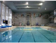 Piscina coperta con grande vasca rettangolare e corsie, illuminata da ampie finestre laterali. Sul fondo si vede una piattaforma per tuffi e grandi murales figurativi sulle pareti, con gradinate per spettatori su un lato.