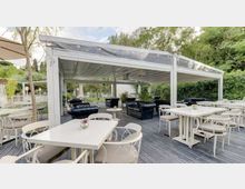 Terrazza all’aperto con pergolato bianco e copertura trasparente, pavimentazione in legno e tavoli bianchi con sedie coordinate. Sul fondo sono presenti divani scuri da lounge e siepi e alberi che circondano l’area.
