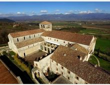 Veduta aerea di un grande complesso monastico in pietra con tetti in coppi e campanile quadrato, disposto attorno a un cortile interno con porticato. L’edificio è circondato da campi e strade di campagna, con una catena montuosa sullo sfondo sotto un cielo sereno.