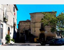 Piazzetta urbana con edifici in muratura intonacata: a sinistra una casa con balconi e persiane, al centro un edificio storico con portale ad arco e piccola loggia. In primo piano si vedono auto parcheggiate e un albero sul lato destro, sotto un cielo sereno.