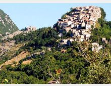 Borgo collinare con case in pietra addossate tra loro, arroccato su un pendio boscoso. Intorno si vedono colline verdi e un paesaggio montano sotto cielo sereno.