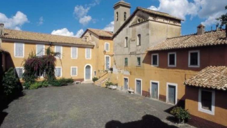 Cortile in pietra di un complesso storico con edifici intonacati color ocra e rosso, finestre con cornici bianche e tetti in coppi. Sullo sfondo si vede una chiesa con campanile e facciate in muratura sotto un cielo azzurro con nuvole.