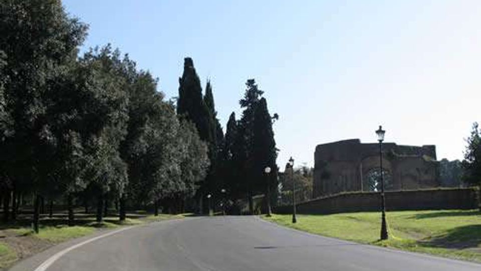 Strada asfaltata ampia e curva che attraversa un’area verde, fiancheggiata da alberi alti e lampioni. Sullo sfondo si vede un edificio in muratura con un muro di recinzione, sotto un cielo limpido.