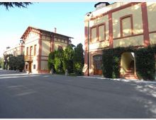 Complesso di edifici in stile villa storica con facciate color crema e cornici rosse, affacciati su un ampio piazzale asfaltato. Sono visibili finestre alte, un portico con scala d’ingresso e siepi e alberi sempreverdi lungo i lati.