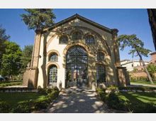 Facciata di un edificio storico con grande ingresso ad arco vetrato centrale e finestre ad arco, raggiunto da un vialetto in pietra. L’edificio è circondato da prato, aiuole e alti alberi in un’area verde.