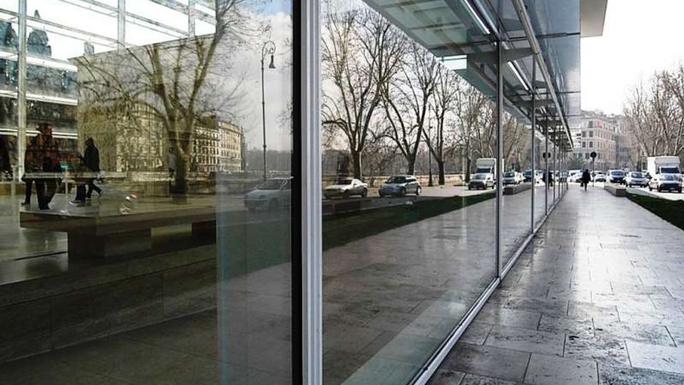 Marciapiede lungo un edificio moderno con grande facciata vetrata riflettente e pensilina in vetro e metallo. Accanto corre una strada con auto e alberi spogli, con edifici sullo sfondo.