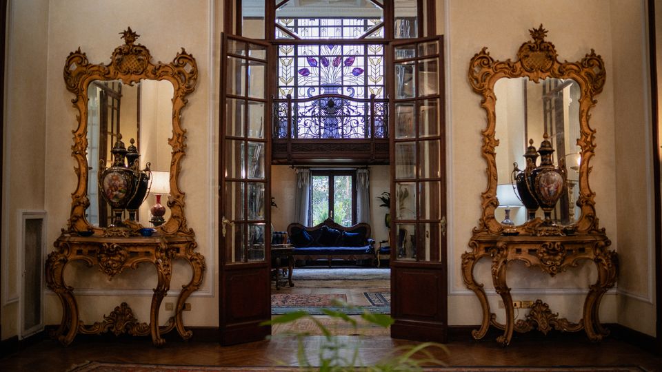 Interno di un elegante ingresso con porte a vetri aperte e grande finestra con vetrata decorata sopra l’architrave. Ai lati, due consolle e specchi barocchi dorati simmetrici; sullo sfondo si intravede un salotto con divano, tappeti e luce naturale dalle finestre.
