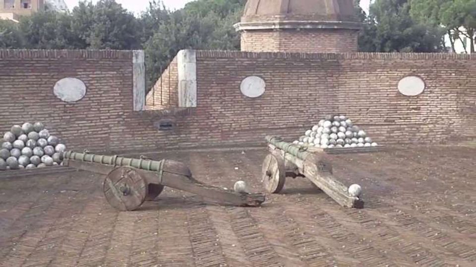 Cortile in mattoni con due carri/cataste in legno che trasportano pile di palle di cannone in metallo. Sullo sfondo si vede un alto muro in laterizio con targhe ovali e una struttura circolare in muratura, circondata da alberi.