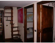 Interno di un’abitazione con scala a giorno in legno scuro e gradini sospesi lungo la parete. Accanto si vedono porte e telai in legno, una porta aperta, e una parete con quadri decorativi e una mappa.