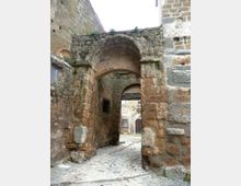 Antico passaggio ad arco in pietra tra muri spessi e irregolari, con tracce di vegetazione tra le fughe. Il pavimento è in lastre di pietra e l’arco conduce a un piccolo cortile con un edificio in muratura e una porta sullo sfondo.