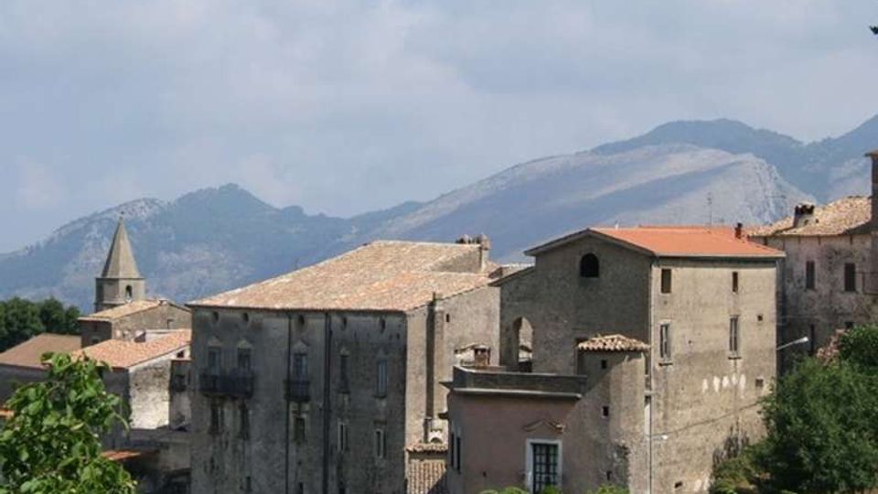 Complesso di edifici in pietra con tetti in coppi, tra cui una chiesa con campanile a guglia, affacciato su una valle verde. Sullo sfondo si vedono montagne e un cielo parzialmente nuvoloso.