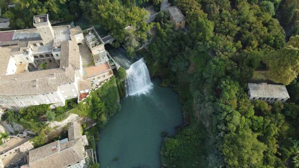 Veduta aerea di un complesso di edifici storici in pietra accanto a una cascata che si tuffa in un bacino d’acqua verde-azzurra. Intorno si estende una fitta area boschiva con sentieri e alcune strutture isolate tra gli alberi.
