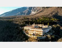 Veduta aerea di un grande complesso monastico fortificato con edifici in pietra e intonaco chiaro, tetti in tegole e un piccolo campanile, racchiuso da mura. Il complesso sorge in una valle boscosa ai piedi di montagne rocciose.