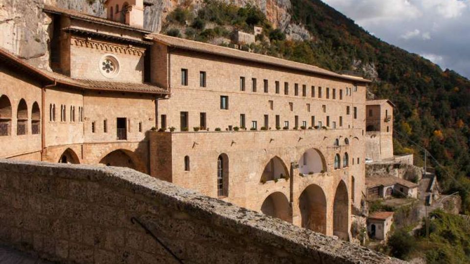 Grande complesso monastico in pietra chiara con arcate e loggiati, costruito a ridosso di una parete rocciosa e disposto su più livelli. Sullo sfondo si vedono il pendio boscoso della valle e il cielo parzialmente nuvoloso.