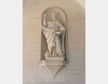 Statua in pietra collocata in una nicchia ad arco su una parete in muratura chiara, con drappeggi scolpiti e base con l’iscrizione “GREGORIO MAGNO”. La figura è su un piedistallo sporgente, con un piccolo elemento decorativo accanto alla base.