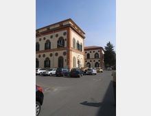 Edificio a più piani in stile storico-industriale con facciata chiara, cornici in mattoni rossi e numerose finestre ad arco e oculi circolari. Davanti si trova un ampio parcheggio asfaltato con auto, con un secondo corpo di fabbrica sullo sfondo e un albero alto sul lato destro.