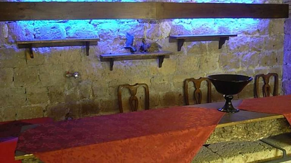 Interno in pietra con volta a botte e parete di blocchi illuminata da luci blu e tre applique circolari. In primo piano un bancone in muratura con inserti in ferro battuto e tovaglia rossa, con sedie e mensole in legno sullo sfondo.