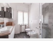 Bagno moderno e luminoso con piastrelle bianche e pavimento grigio scuro, mobile lavabo con piano in legno e pensili scuri. Sul fondo c’è una finestra con tenda e radiatore, mentre a destra si vedono WC, bidet e una doccia con box in vetro.