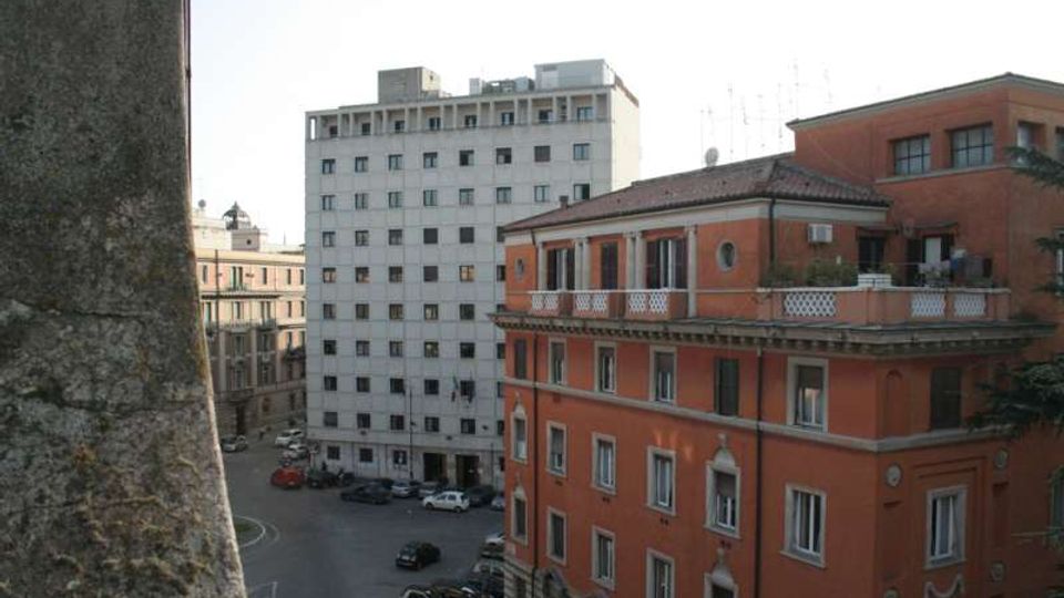 Veduta urbana con palazzo moderno bianco a più piani e edificio storico in mattoni rossi con terrazza e balconate; in basso si vede un incrocio stradale con auto parcheggiate tra altri edifici.