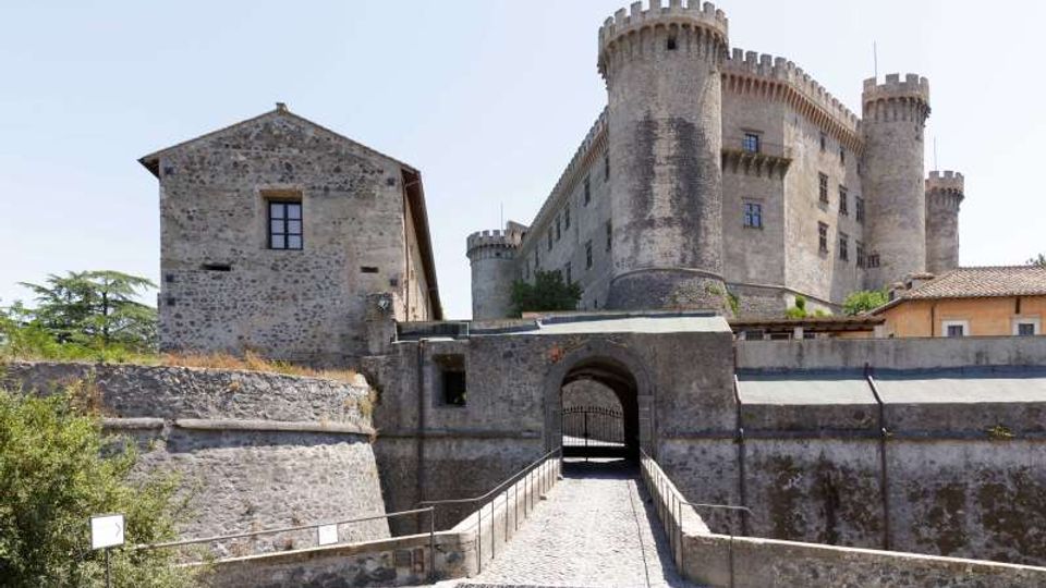 Castello in pietra con torri cilindriche merlate e mura fortificate, visto dall’ingresso con arco e ponte pedonale in ciottoli che conduce al portale. Sullo sfondo cielo sereno e vegetazione bassa ai lati delle fortificazioni.