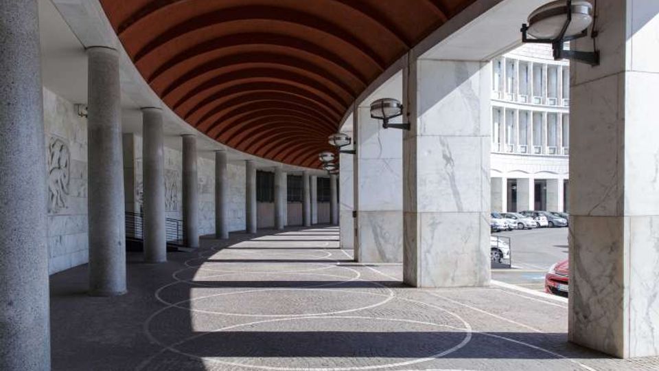 Portico coperto lungo e curvilineo con arcate e soffitto in legno, sostenuto da colonne e pilastri rivestiti in marmo con lampade a parete. Pavimentazione in pietra con motivi circolari e vista laterale su una strada con auto parcheggiate e un edificio moderno sullo sfondo.