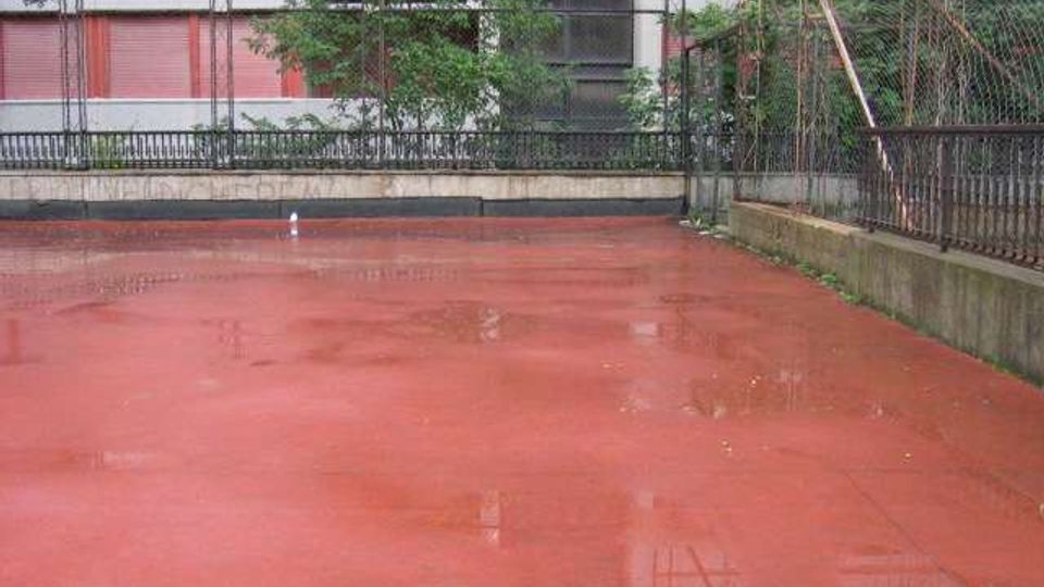 Campo sportivo all’aperto con pavimentazione rossa bagnata dalla pioggia e pozzanghere, circondato da recinzioni metalliche. Sullo sfondo si vedono alberi e un edificio con facciata a pannelli bianchi e rossi.