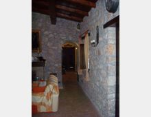Interno rustico con pareti in pietra a vista e soffitto con travi in legno, pavimento in cotto e corridoio che conduce a una porta ad arco. Sulla destra ci sono una finestra con tende leggere e una applique in ferro battuto; a sinistra si intravede un divano vicino a un camino.