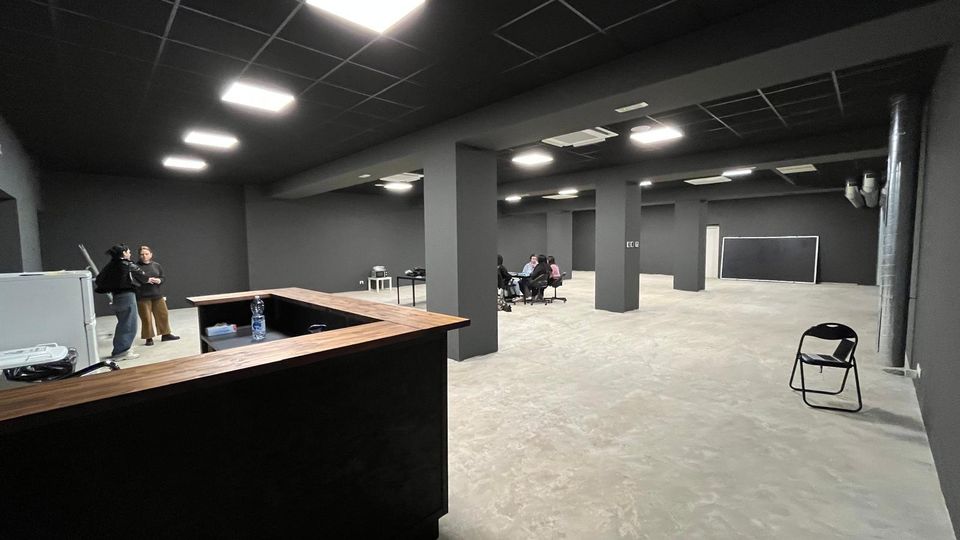 Ampio spazio interno open space in stile industriale, con pareti grigio scuro, colonne portanti e soffitto a griglia con pannelli luminosi. Pavimento in cemento e arredi essenziali, tra cui un bancone in legno e alcune sedie, con una grande lavagna a parete sul fondo.