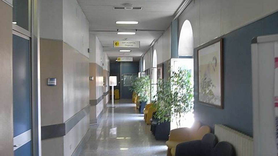 Corridoio interno di un ospedale con pavimento lucido, pareti bicolori e illuminazione a soffitto, con segnaletica sospesa che indica “Sala chirurgica”. Lungo il lato destro ci sono sedie d’attesa, piante in vaso e finestre ad arco che illuminano il passaggio.